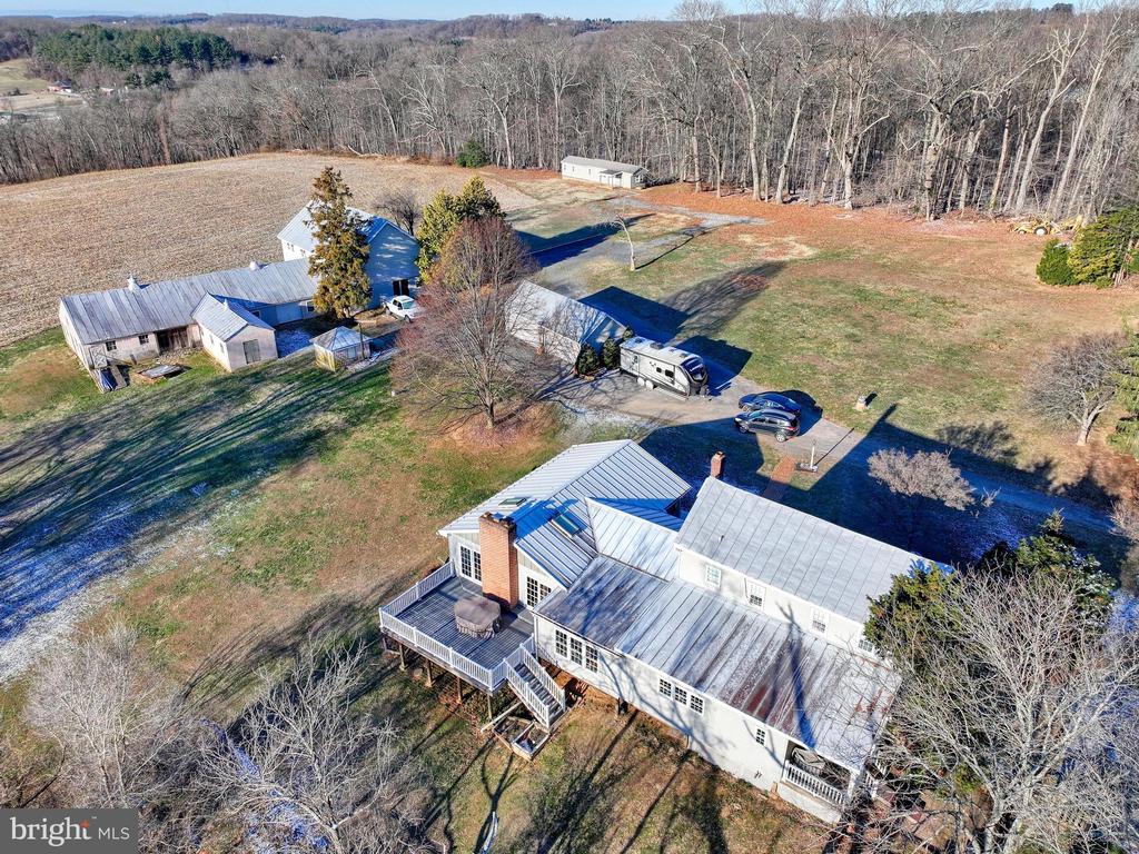 Aerial view of a rural home with a broad deck, outbuildings, and open fields bordered by woods.