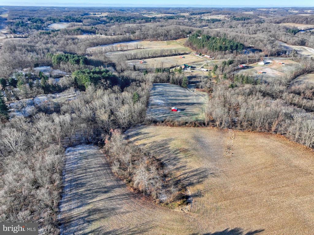 Aerial view of open fields bordered by leafless woods, with scattered rural homes in the distance.
