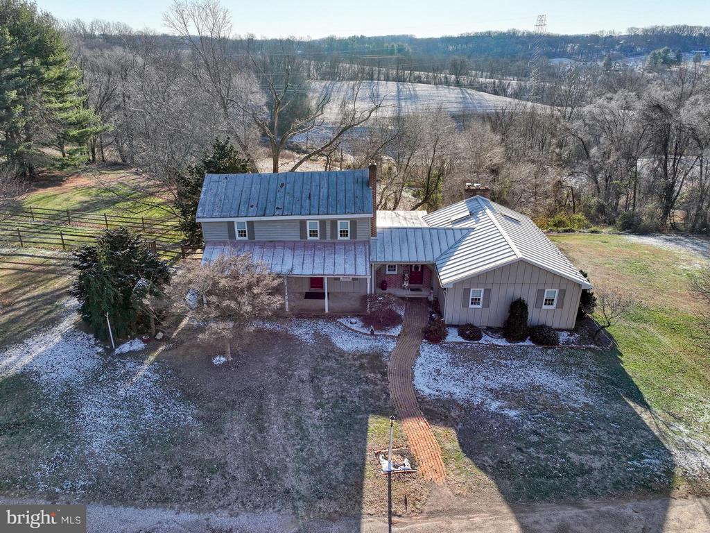 Aerial view of a gray home with metal roofs, a curved brick walkway, and wooded hills beyond.