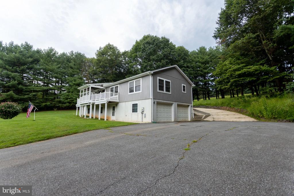 Gray-and-white home with a raised covered porch, two garages, a broad driveway, and wooded surroundings.