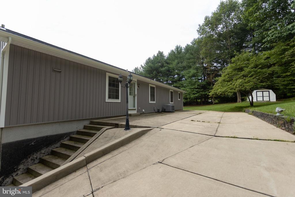 Gray single-story home beside a broad concrete driveway, with entry steps, a white shed, and mature trees.