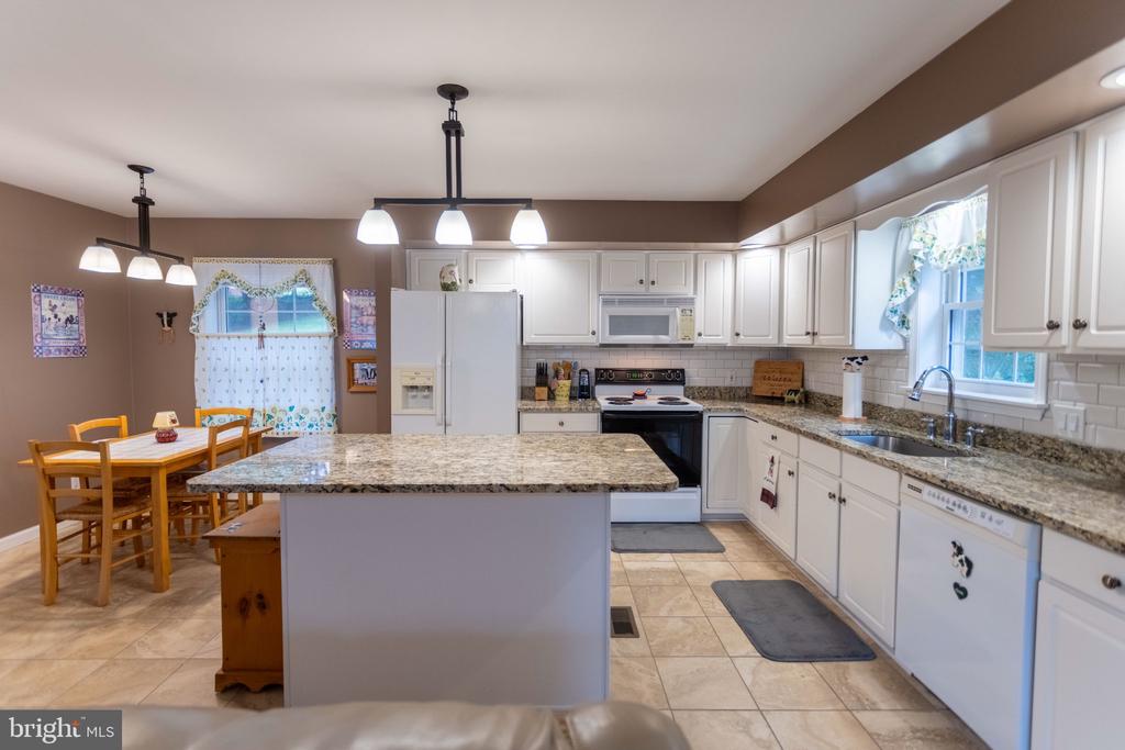 Kitchen with white cabinetry, a granite island, stainless appliances, and a dining table by a curtained window.