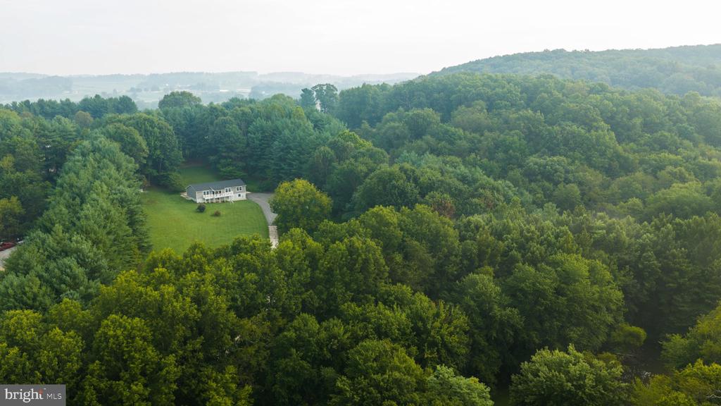 Aerial view of a house in a forest clearing, with a long driveway and wooded hills beyond.