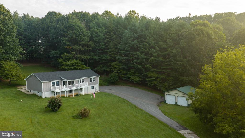 Aerial view of a gray home with a raised deck, detached garage, and wooded surroundings.