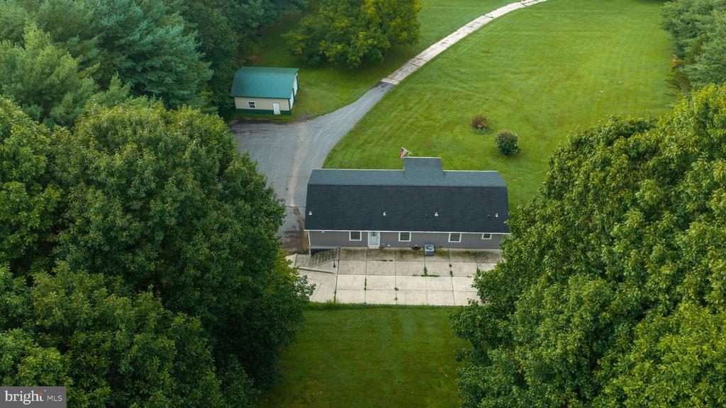 Aerial view of a long building with a paved forecourt, lawn, trees, and detached outbuilding.