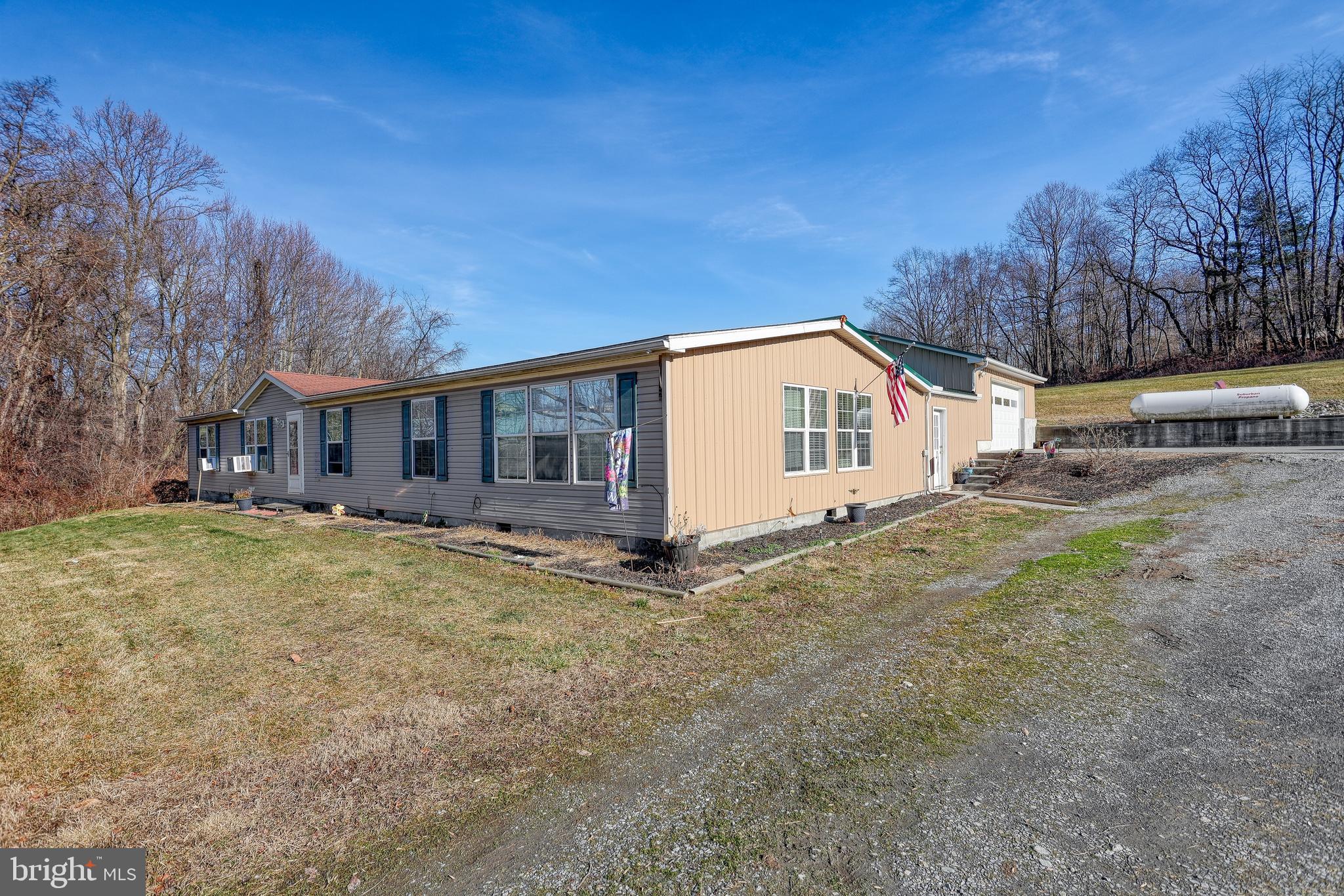 Single-story home with tan and gray siding, an attached garage, gravel driveway, and wooded surroundings.