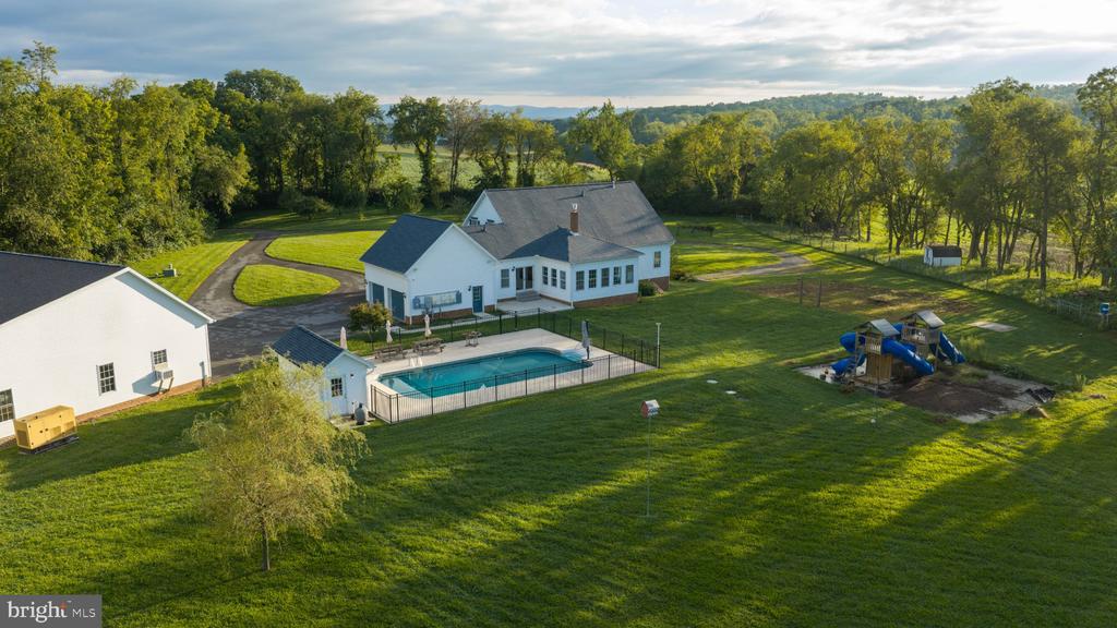 Aerial view of a white home with a fenced pool, detached outbuilding, and playground on a spacious lawn.