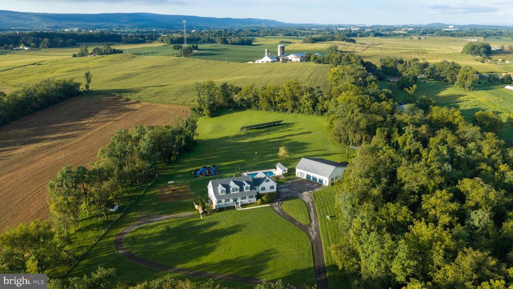 Aerial view of a white home and detached outbuilding amid landscaped grounds, farmland, and woods.