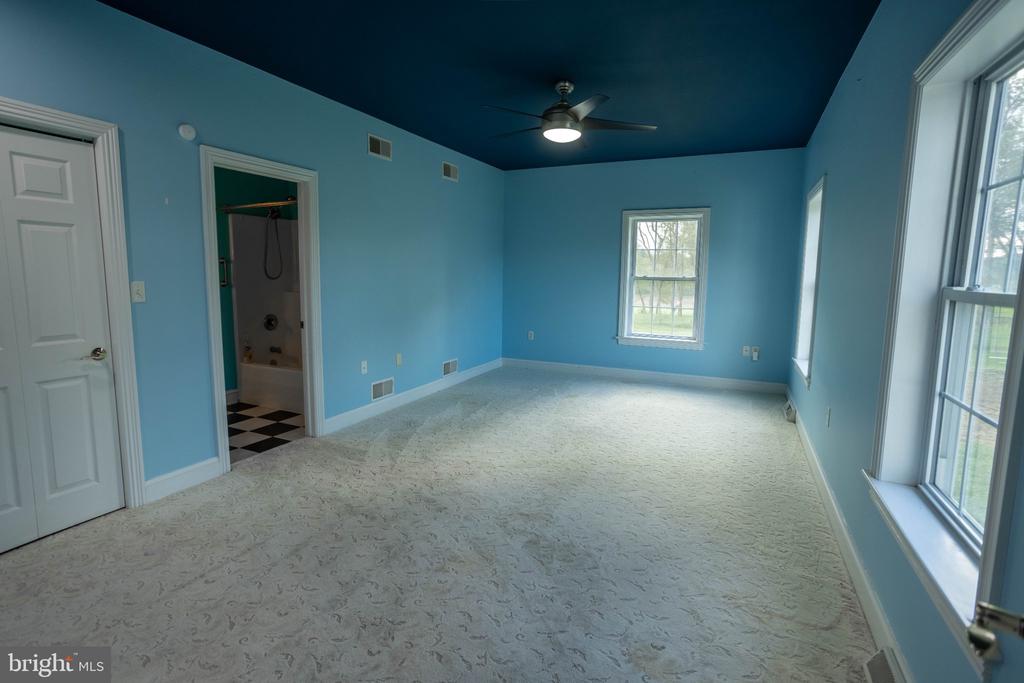 Empty blue bedroom with a ceiling fan, carpet, several windows, and an open bathroom doorway.