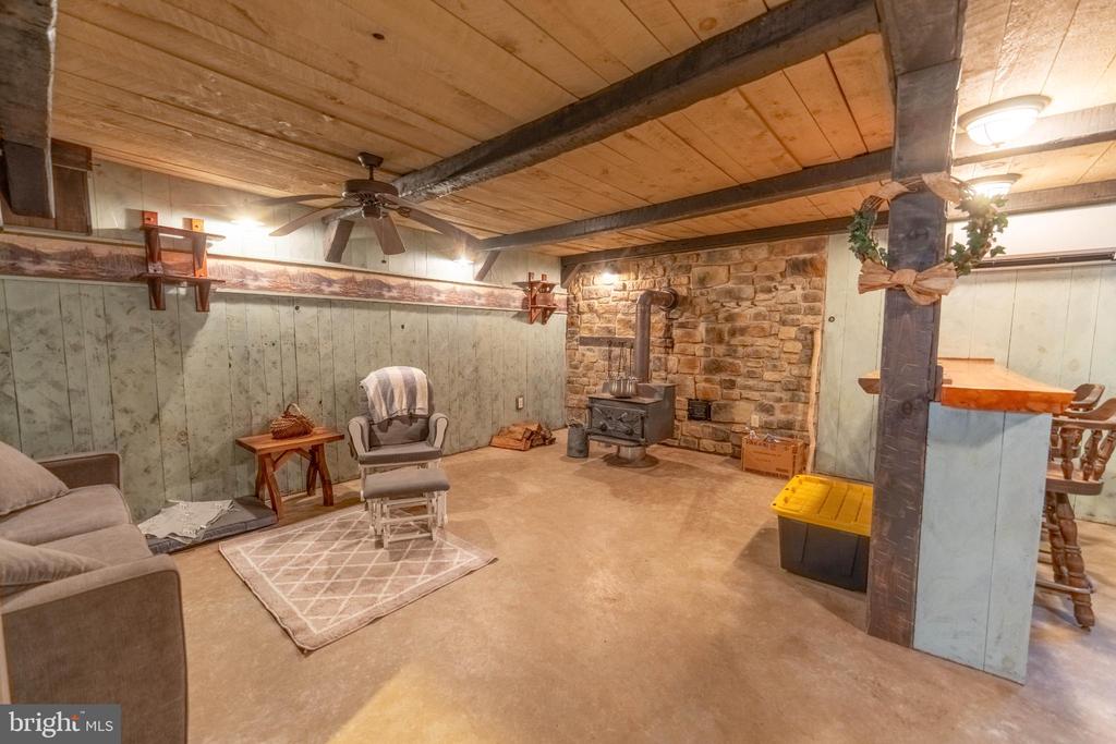 Basement recreation room with exposed wood beams, a stone fireplace, and a freestanding wood stove.