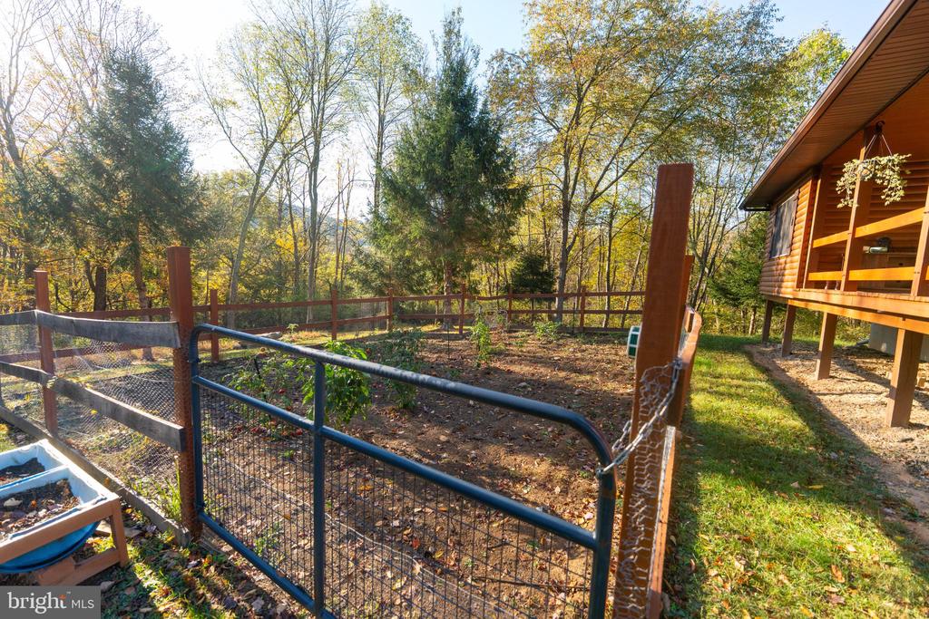 Fenced garden beside a raised log-sided cabin, surrounded by autumn trees.