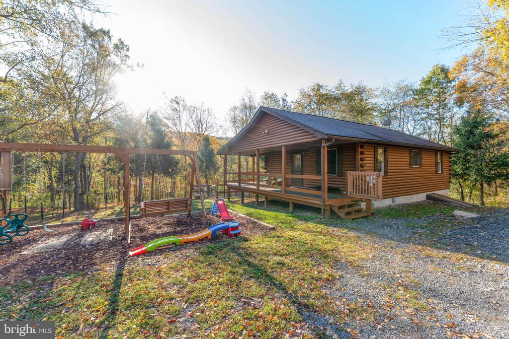Log-sided cabin with a covered porch and children’s play equipment in a wooded autumn yard.