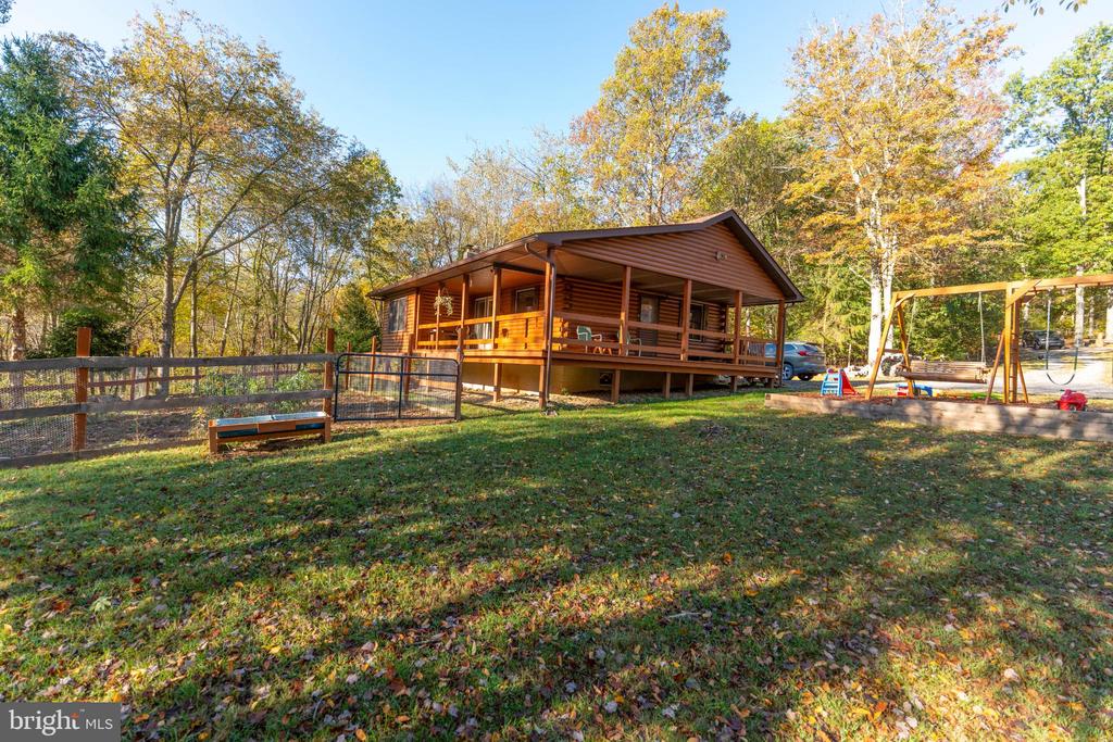 Wood-sided cabin with a covered porch, fenced yard, and swing set among autumn trees.