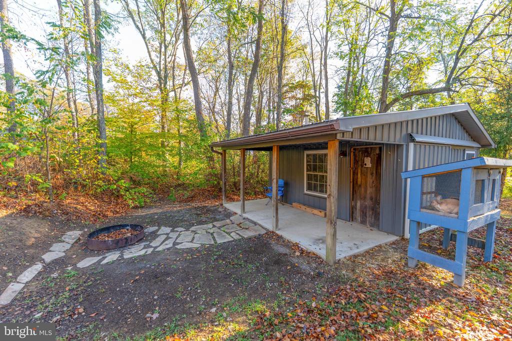Gray outbuilding with a covered porch, stone path, and fire pit in a wooded autumn yard.