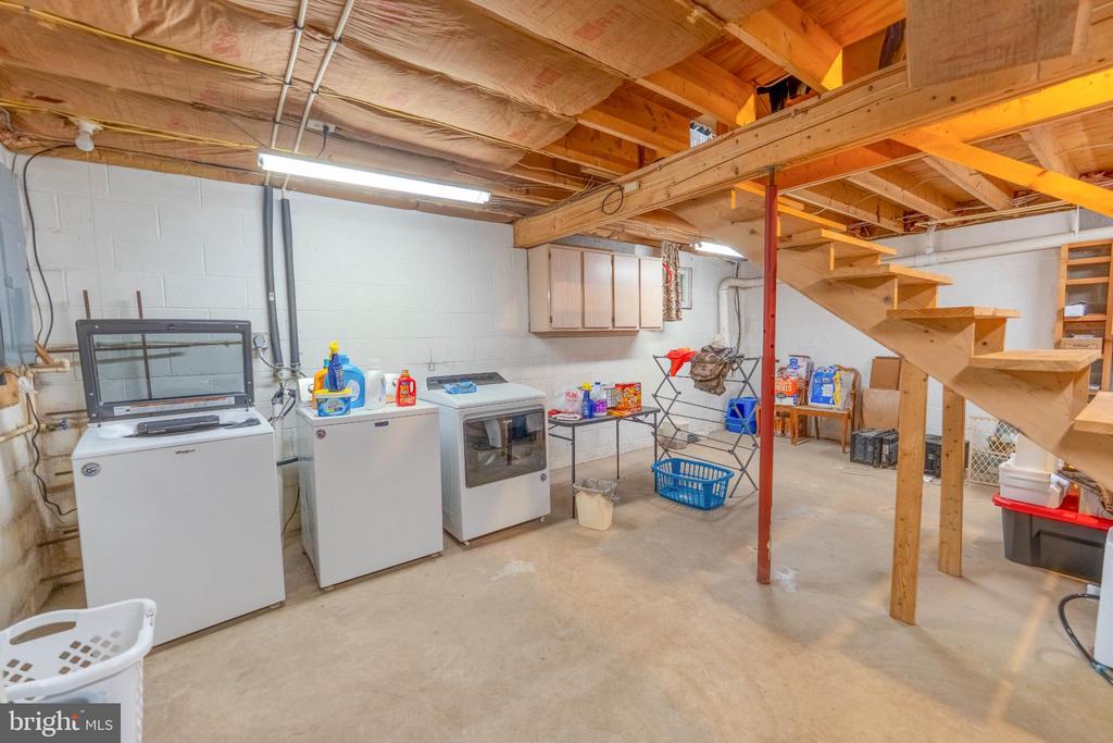 Basement laundry area with washer and dryer beneath exposed beams and beside open wooden stairs.
