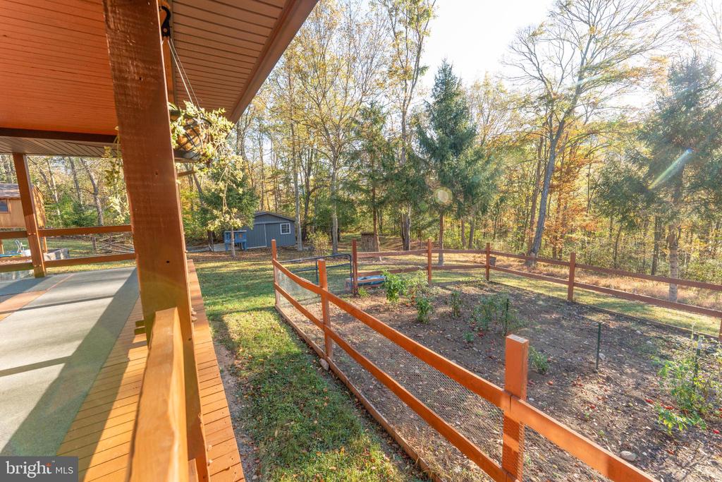 Fenced garden and yard beside a covered wooden porch, with a shed and autumn woods beyond.