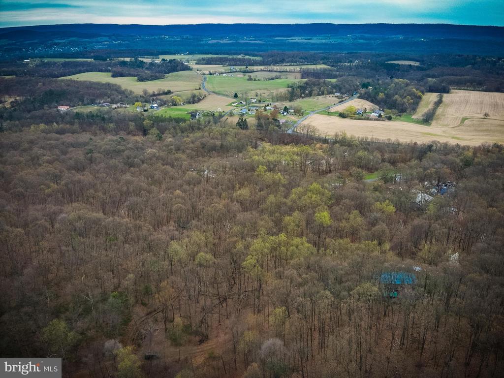 Aerial view of wooded land with a blue-roofed structure, farmland, and a distant ridge.