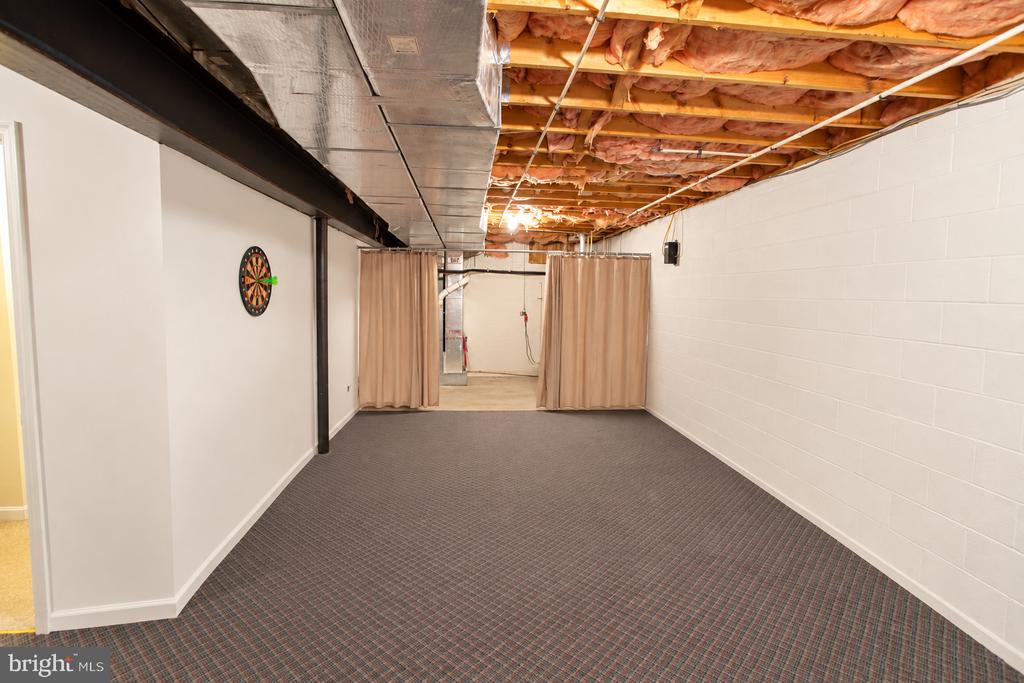 Carpeted basement recreation room with exposed ceiling, beige curtains, and a dartboard.