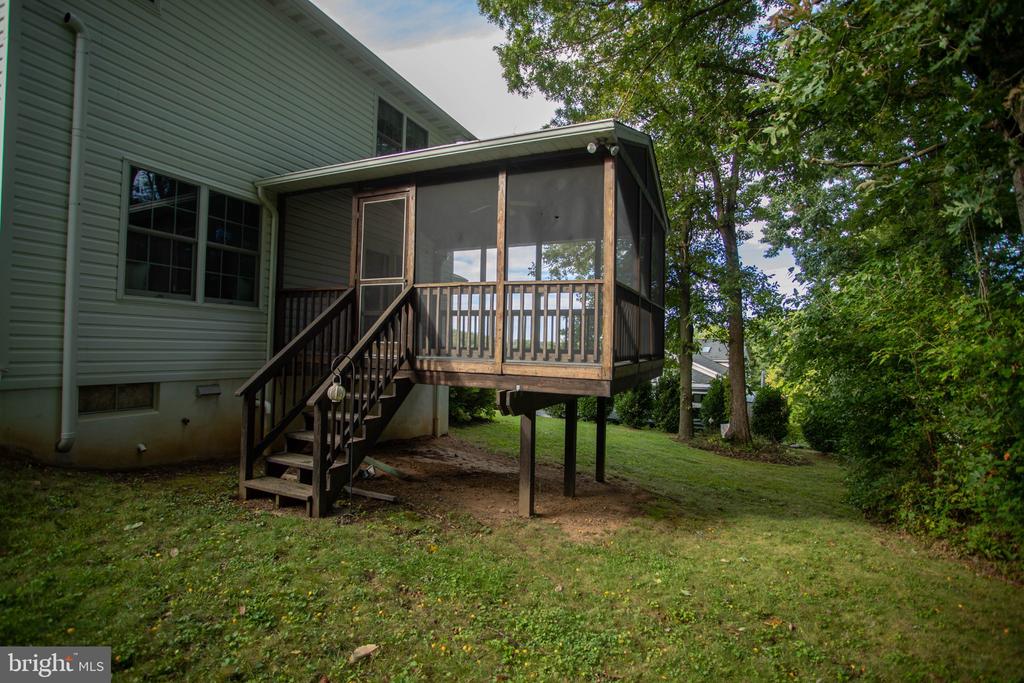 Gray-sided house with an elevated screened porch and wooden stairs beside a grassy, tree-lined yard.