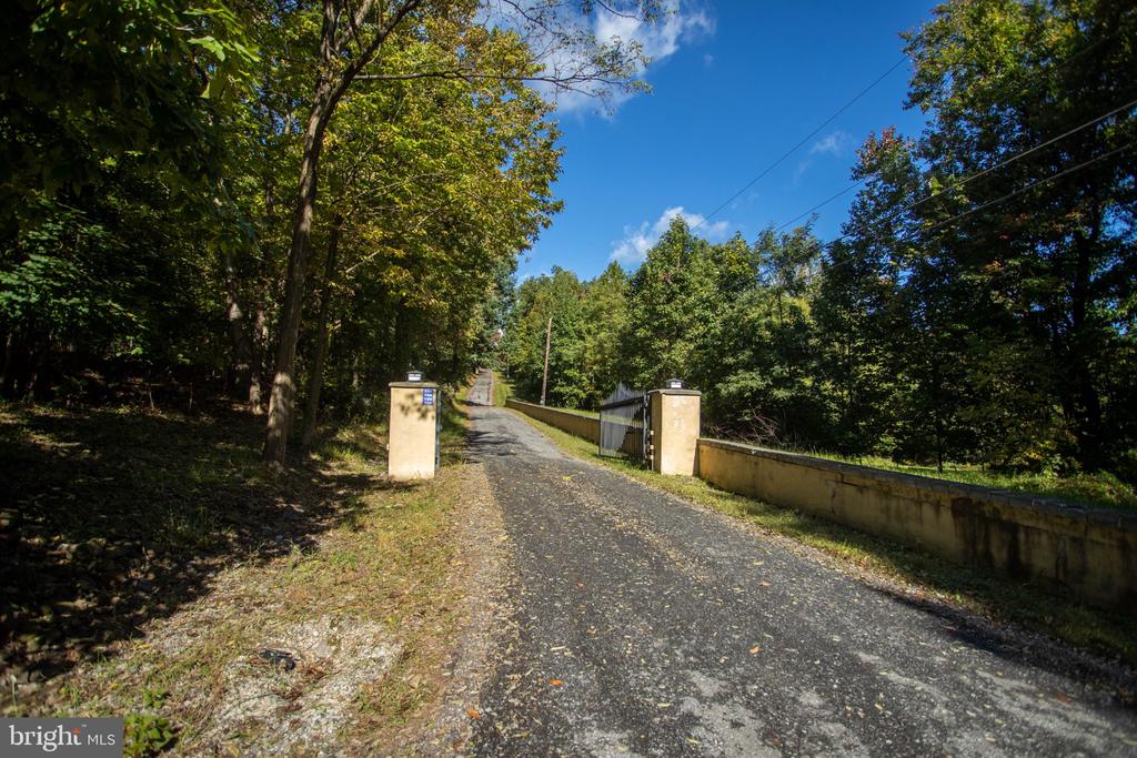 Gravel driveway entrance with stone pillars and a low wall, surrounded by trees.