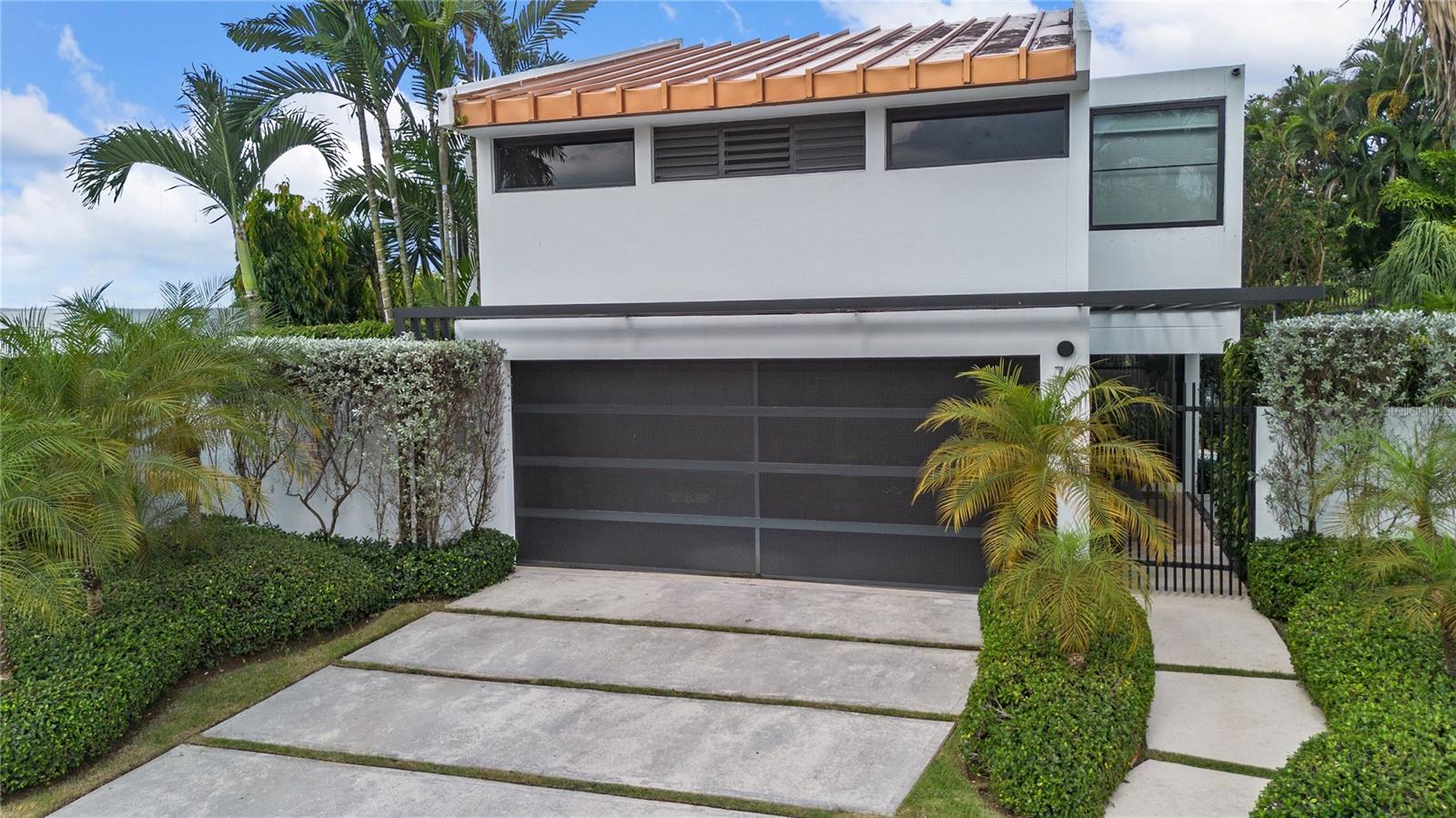 Modern white two-story house with a copper-toned roof, dark garage door, and tropical landscaping.