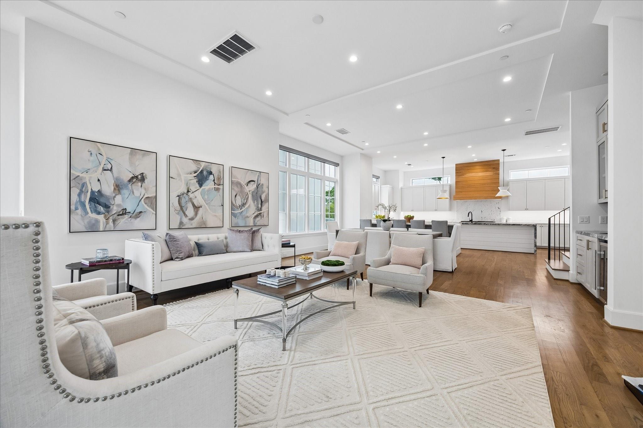 Staged living room opening to a white kitchen, with abstract art, large windows, and a wood-paneled range hood.