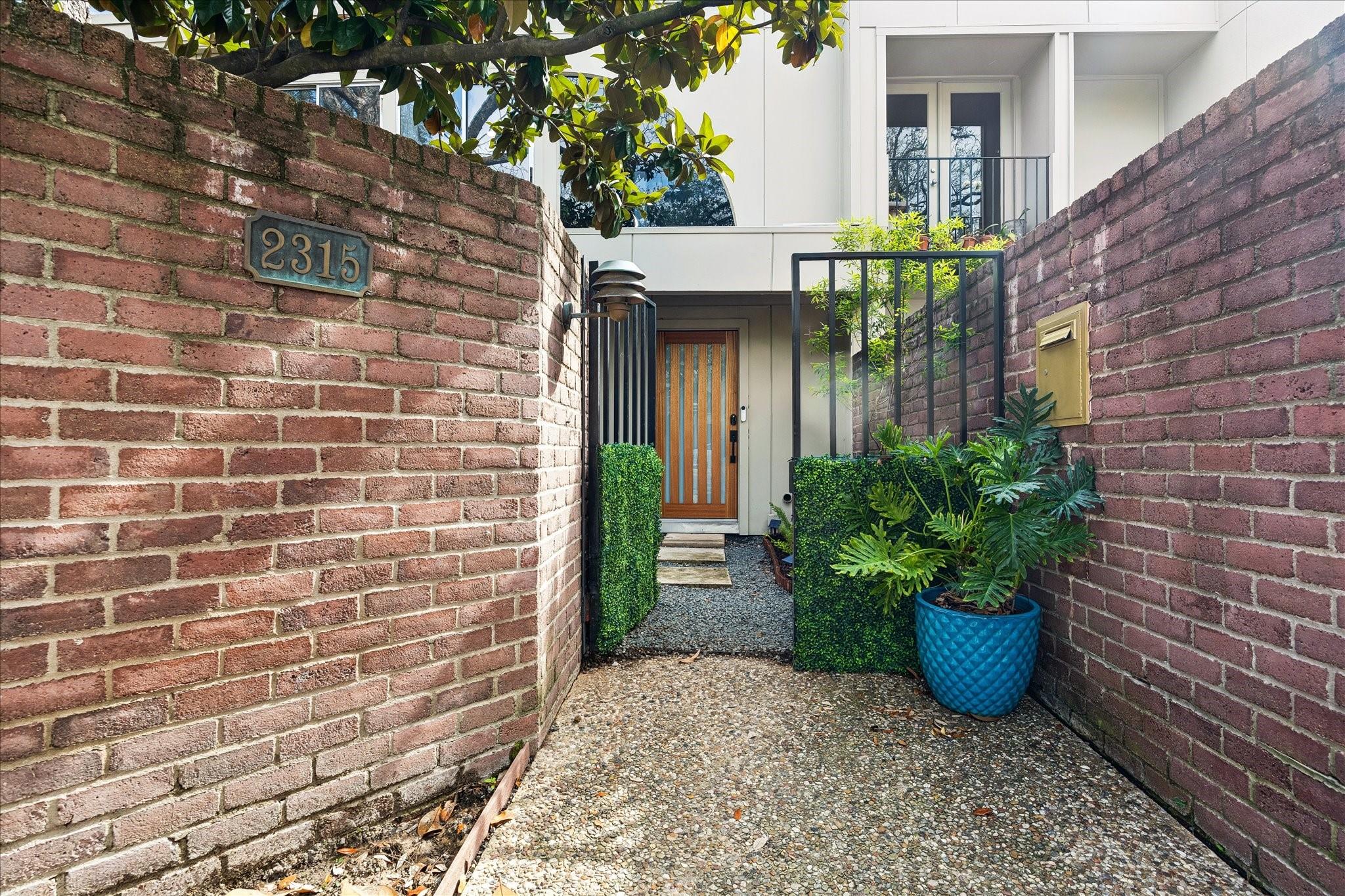 Home entrance framed by brick walls, with a wood-and-glass door and leafy plants.