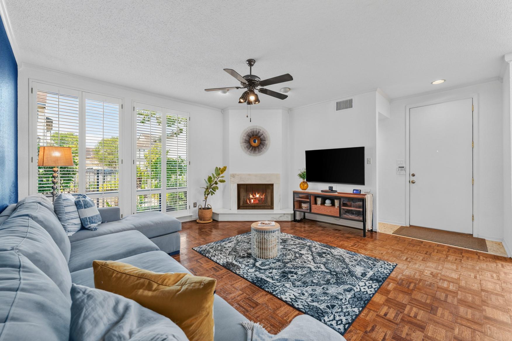 Living room with a corner fireplace, gray sectional sofa, parquet floor, and large shuttered windows.
