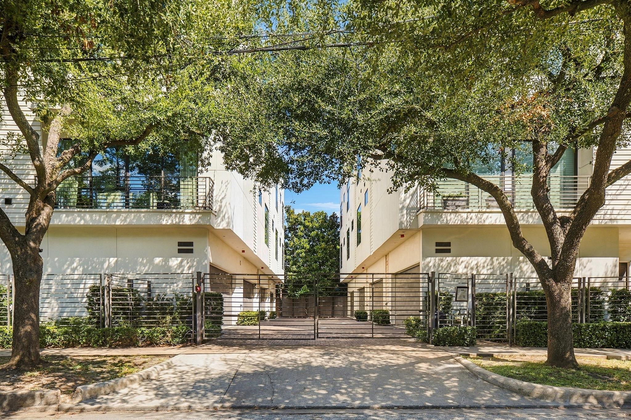 Modern residential building entrance with balconies, metal gate, and large shade trees.