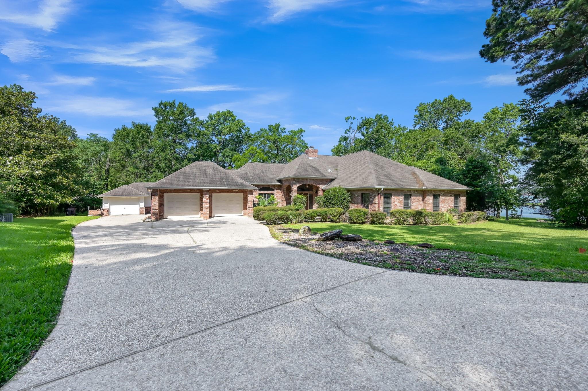Red-brick home with a three-car garage, broad driveway, and water visible through trees.