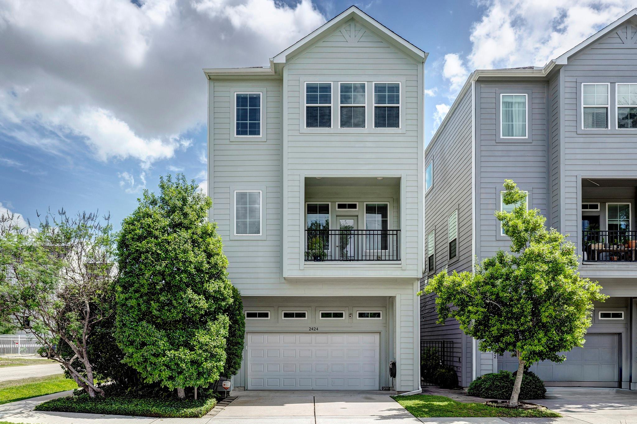 Three-story light gray townhouse with a recessed balcony, garage, and landscaped front yard.