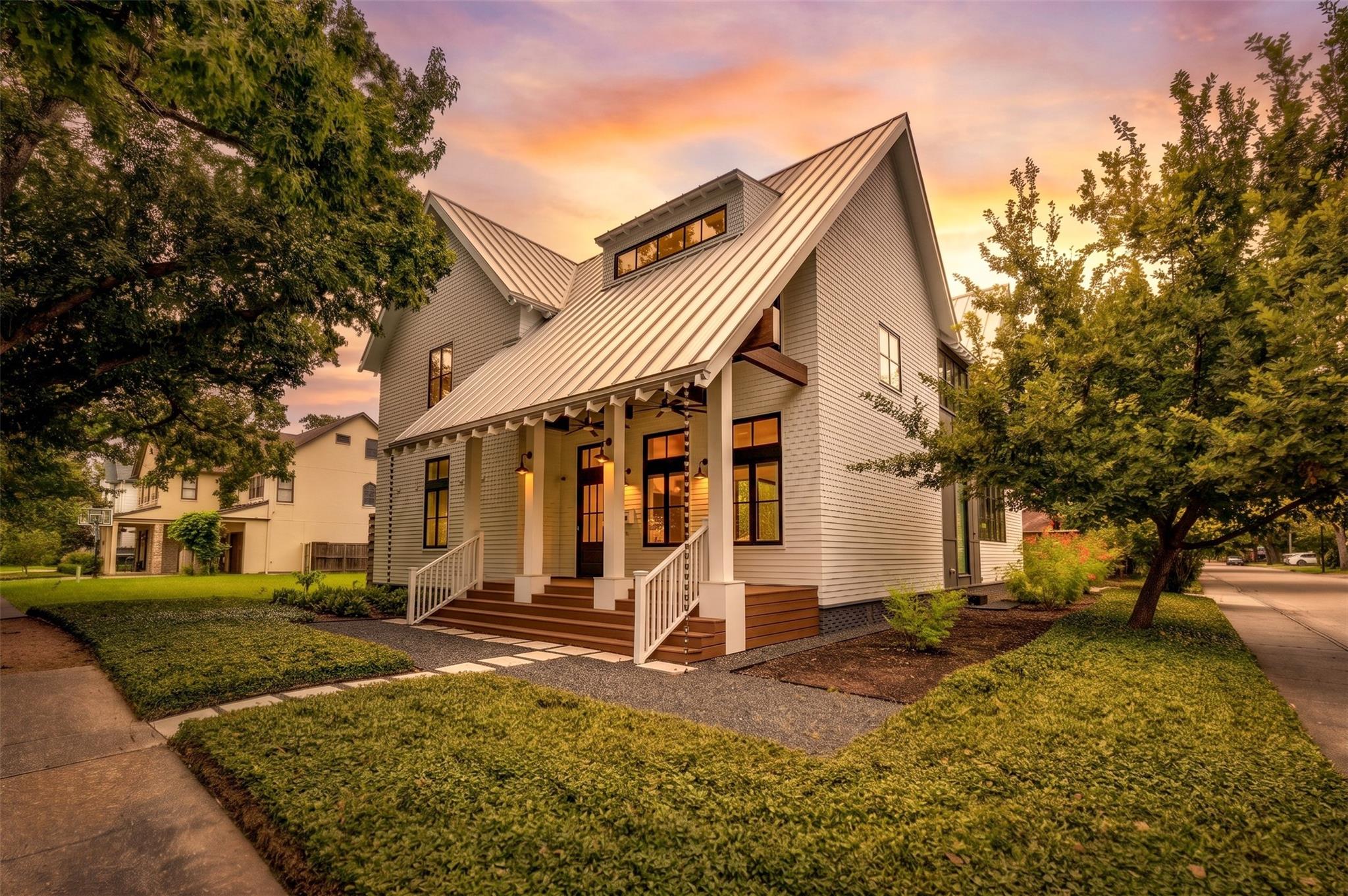White two-story house with a steep metal roof and broad porch, framed by mature trees.