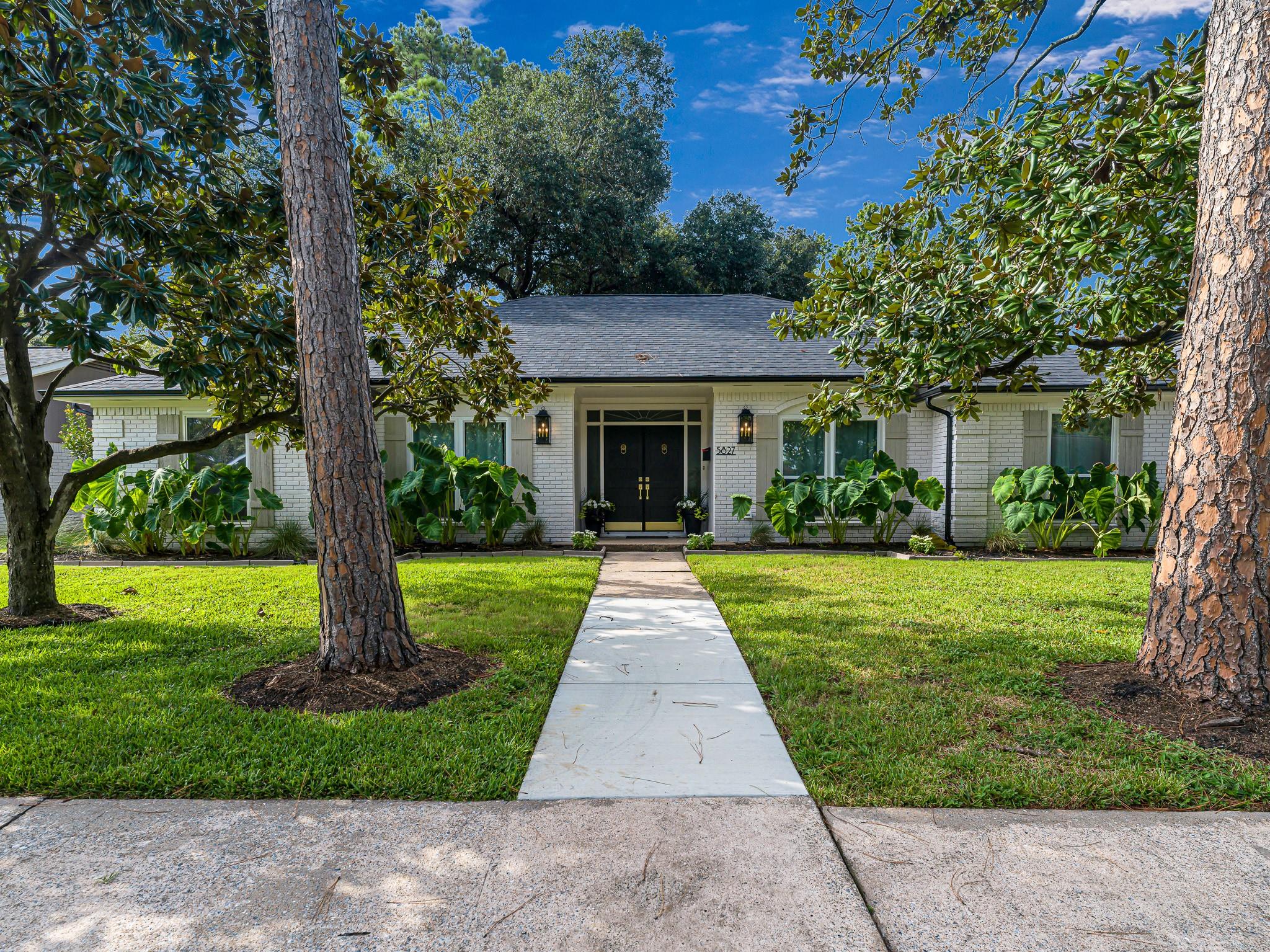 White brick home with black double doors, a landscaped lawn, and a walkway framed by mature trees.