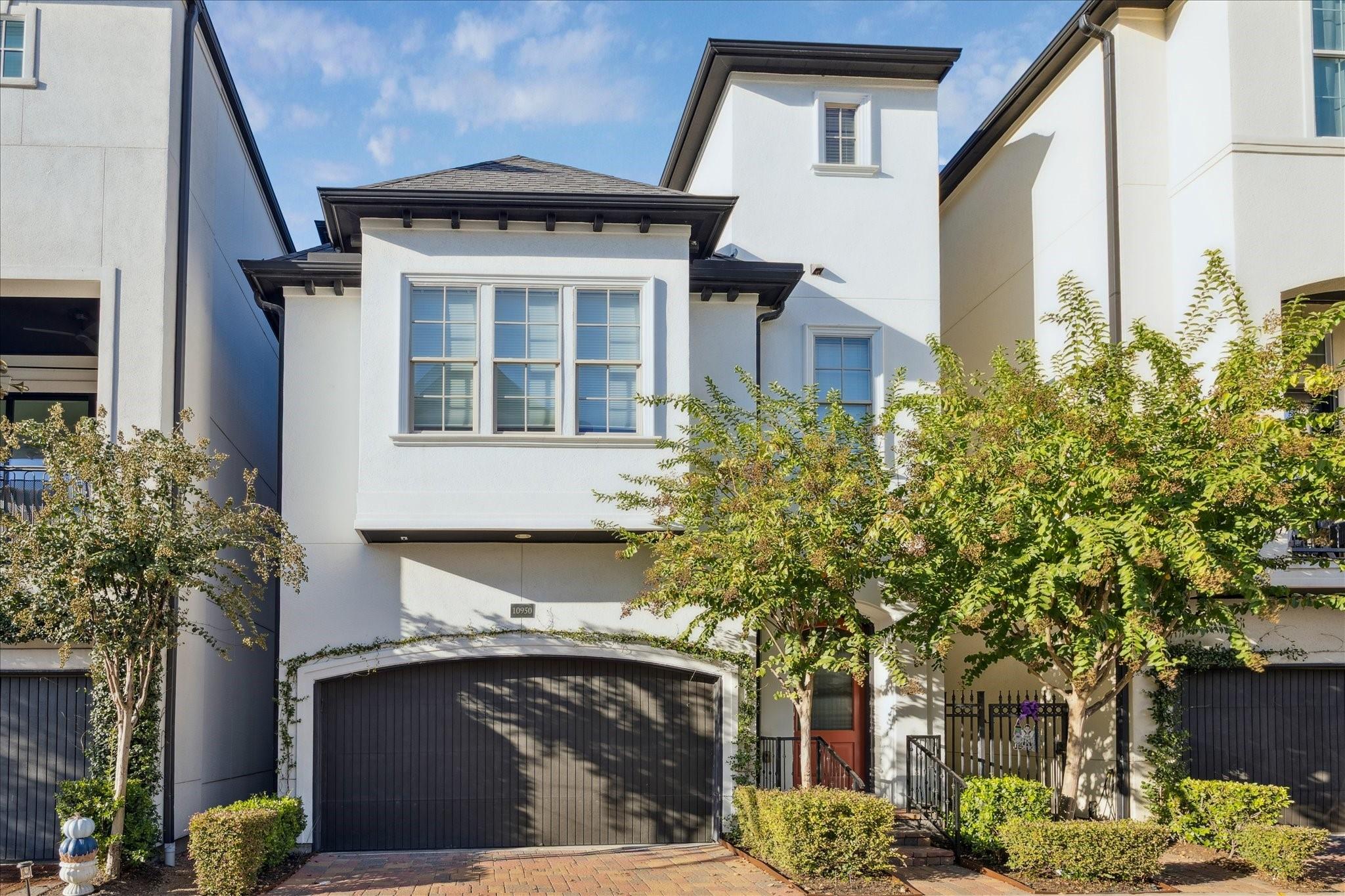 White multi-story townhouse with a dark garage door, front entry, and leafy trees.