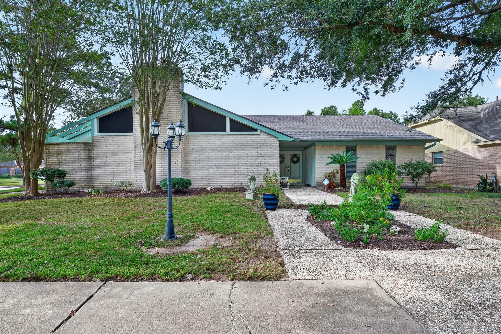 Single-story brick house with teal roof trim, mature shade trees, and a gravel path to the entry.