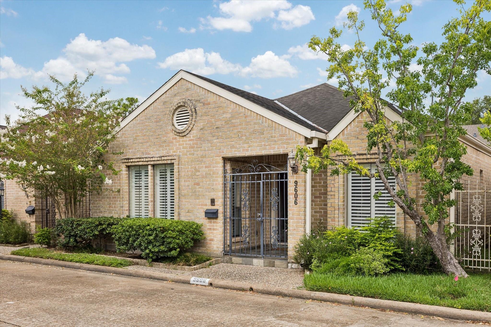 Tan brick home with an ornate metal entry gate, plantation shutters, and landscaped front yard.