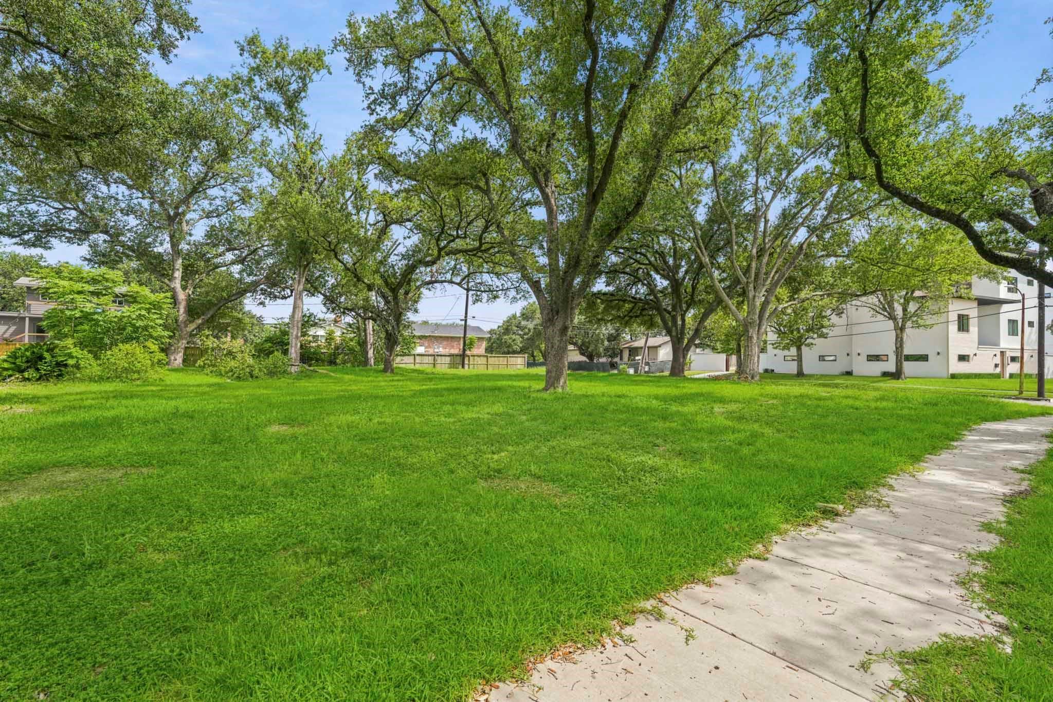 Tree-shaded grassy lot beside a sidewalk, with neighboring homes in the background.