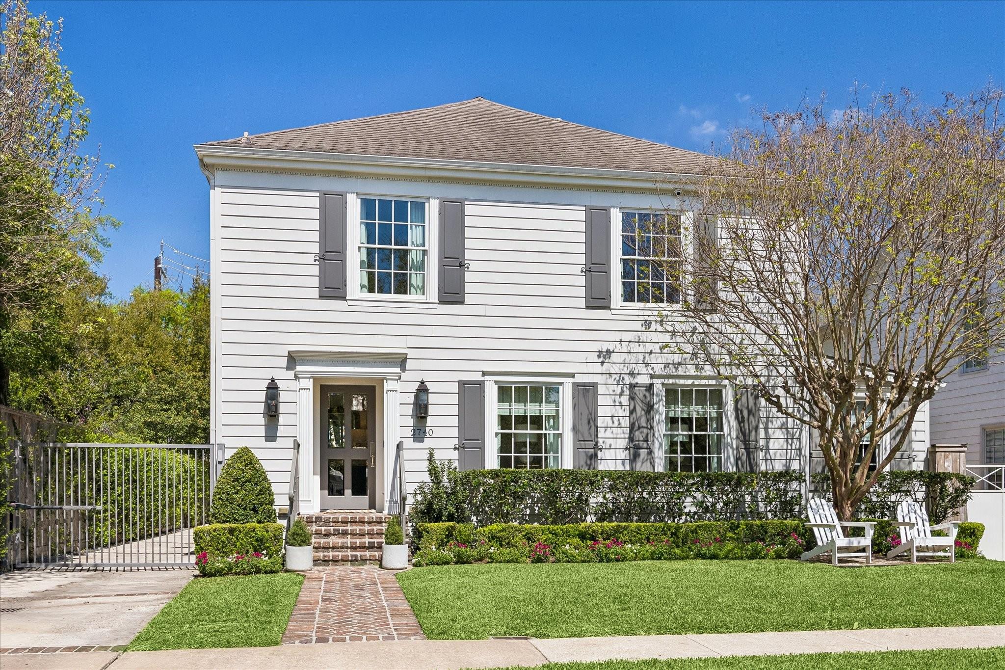 White two-story house with dark shutters, brick walkway, manicured lawn, and Adirondack chairs.