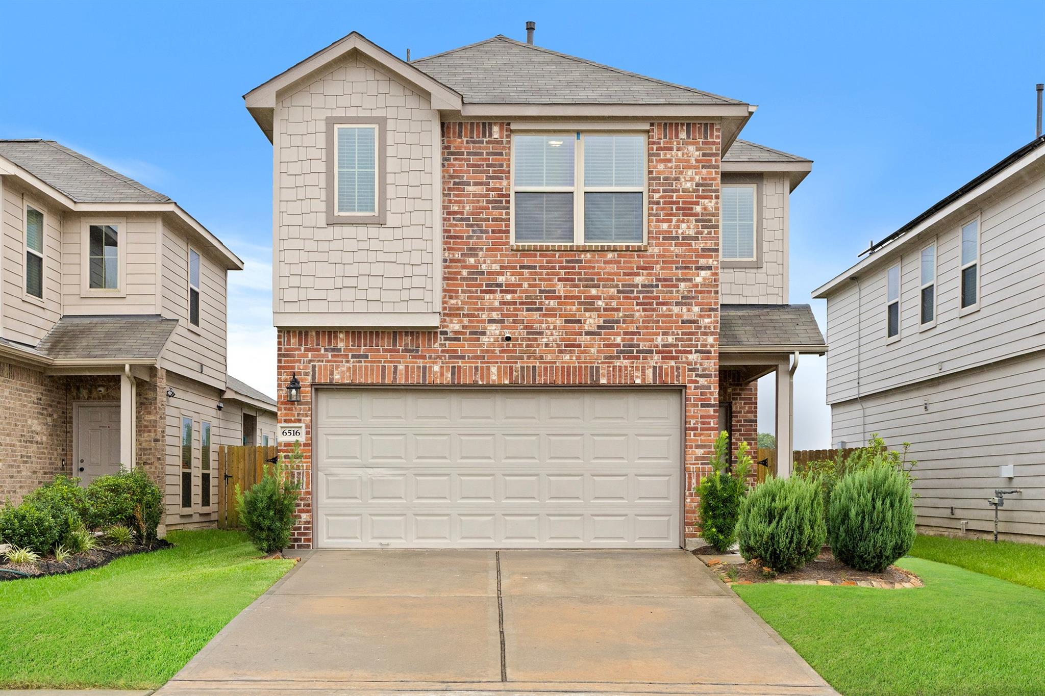 Two-story brick house with a double garage, landscaped shrubs, and a wide concrete driveway.