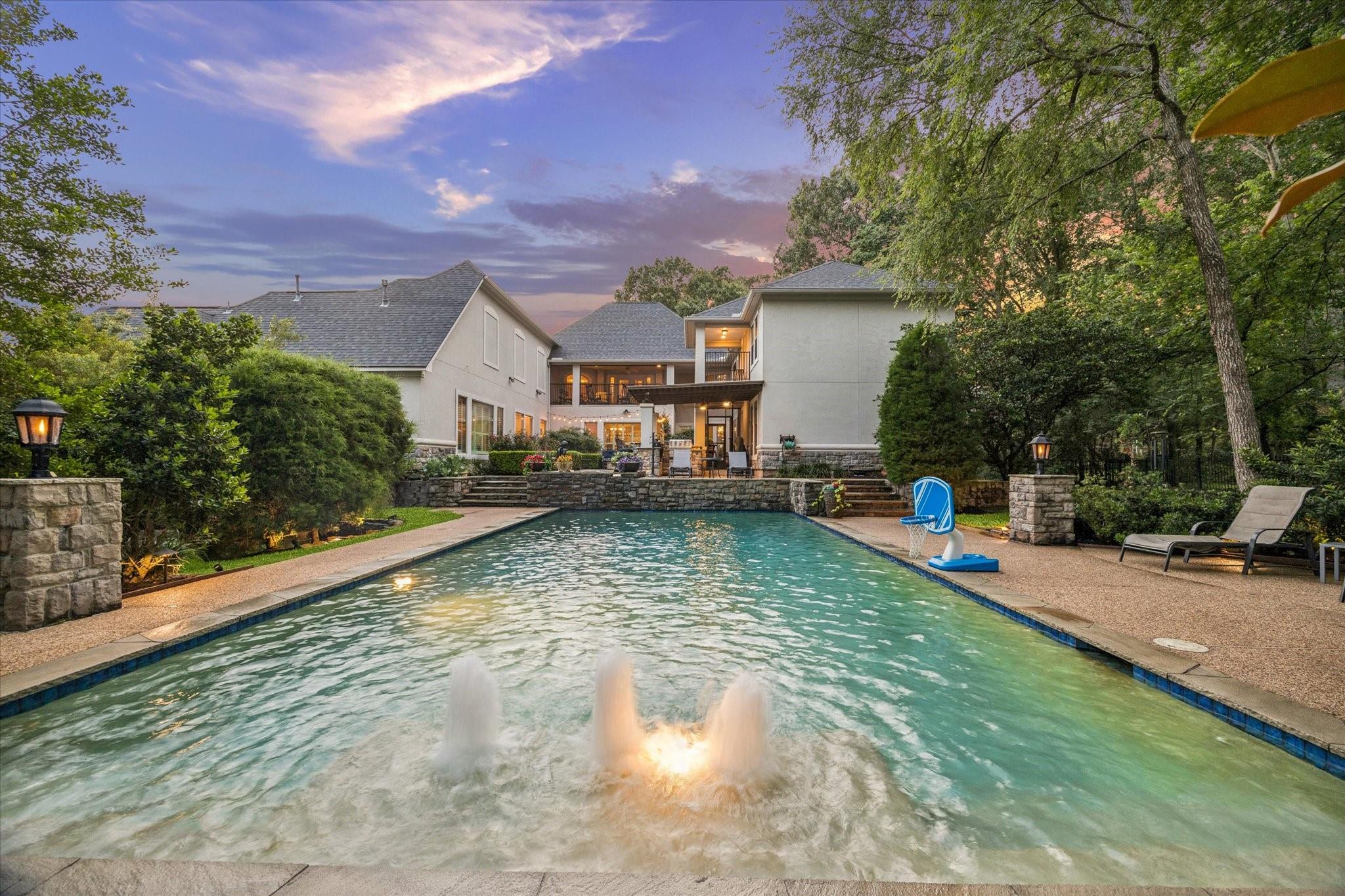 Back of a large house beyond a swimming pool with a fountain, patio seating, and mature trees at dusk.