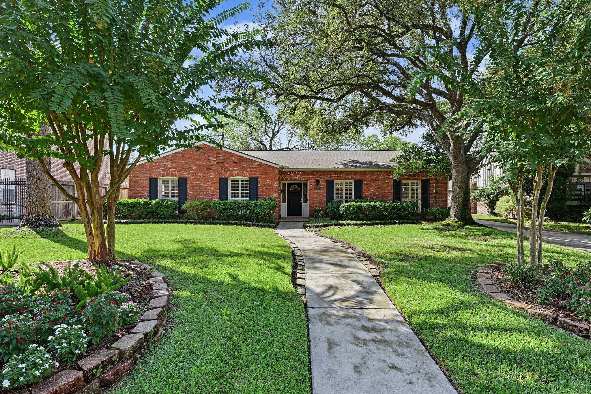 Single-story brick house with dark shutters, a curved walkway, landscaped lawn, and mature trees.