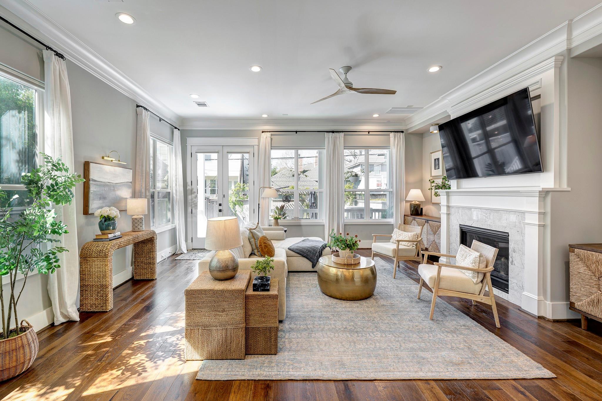 Staged living room with hardwood floors, large windows, cream seating, and a marble fireplace.