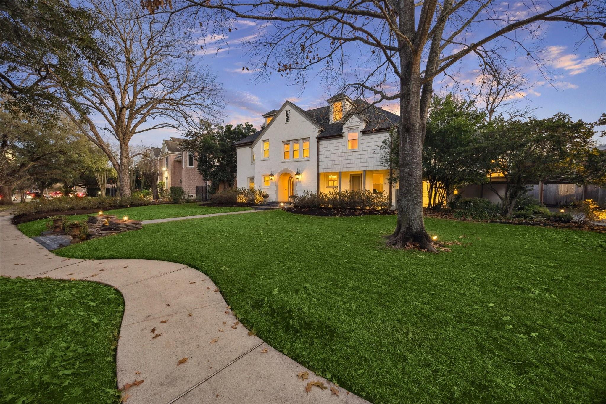 Two-story white house at dusk with glowing windows, mature trees, and a curved walkway across the lawn.