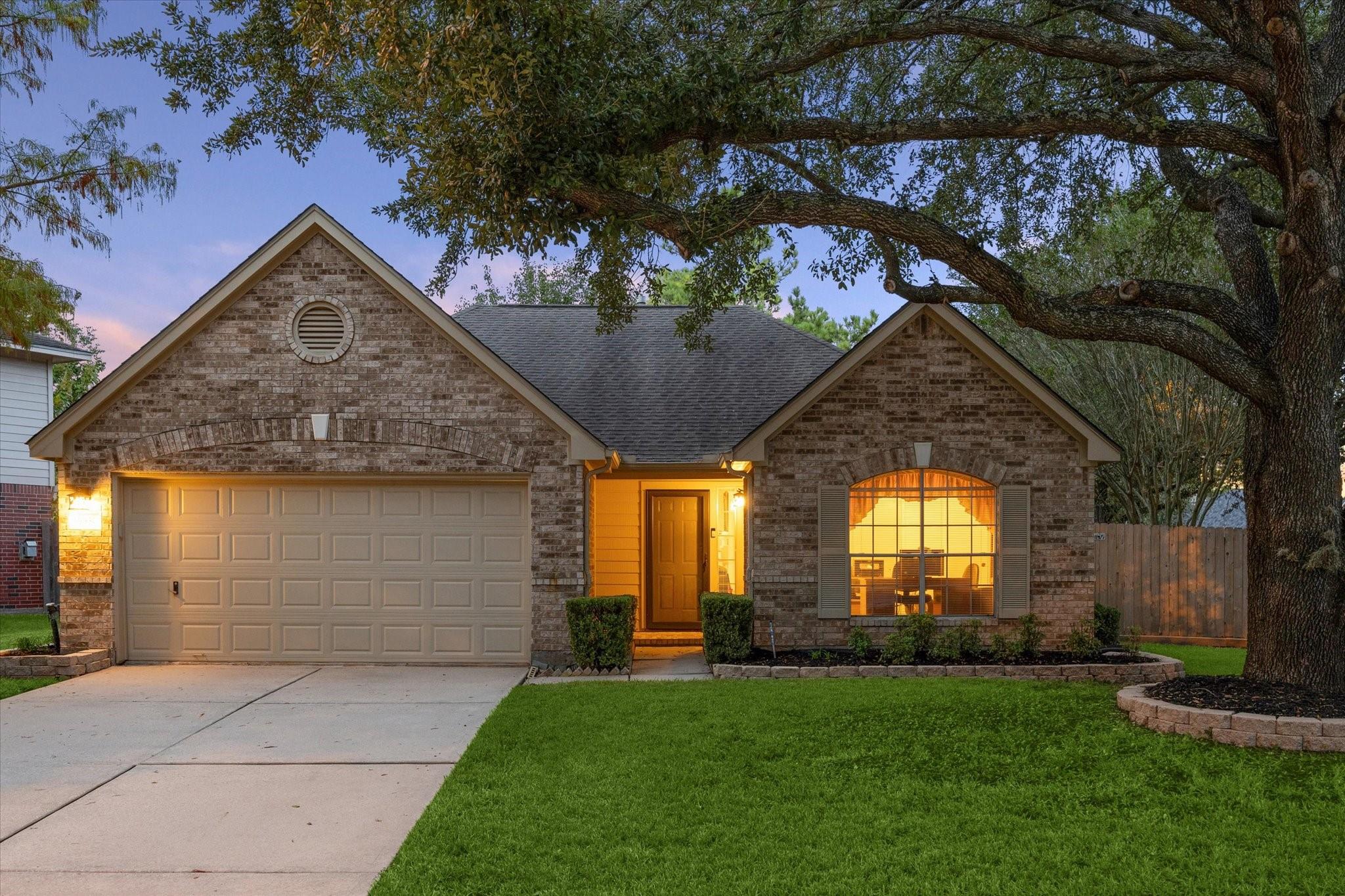 Brick single-story home at dusk with a lit entry and garage beneath a large oak tree.