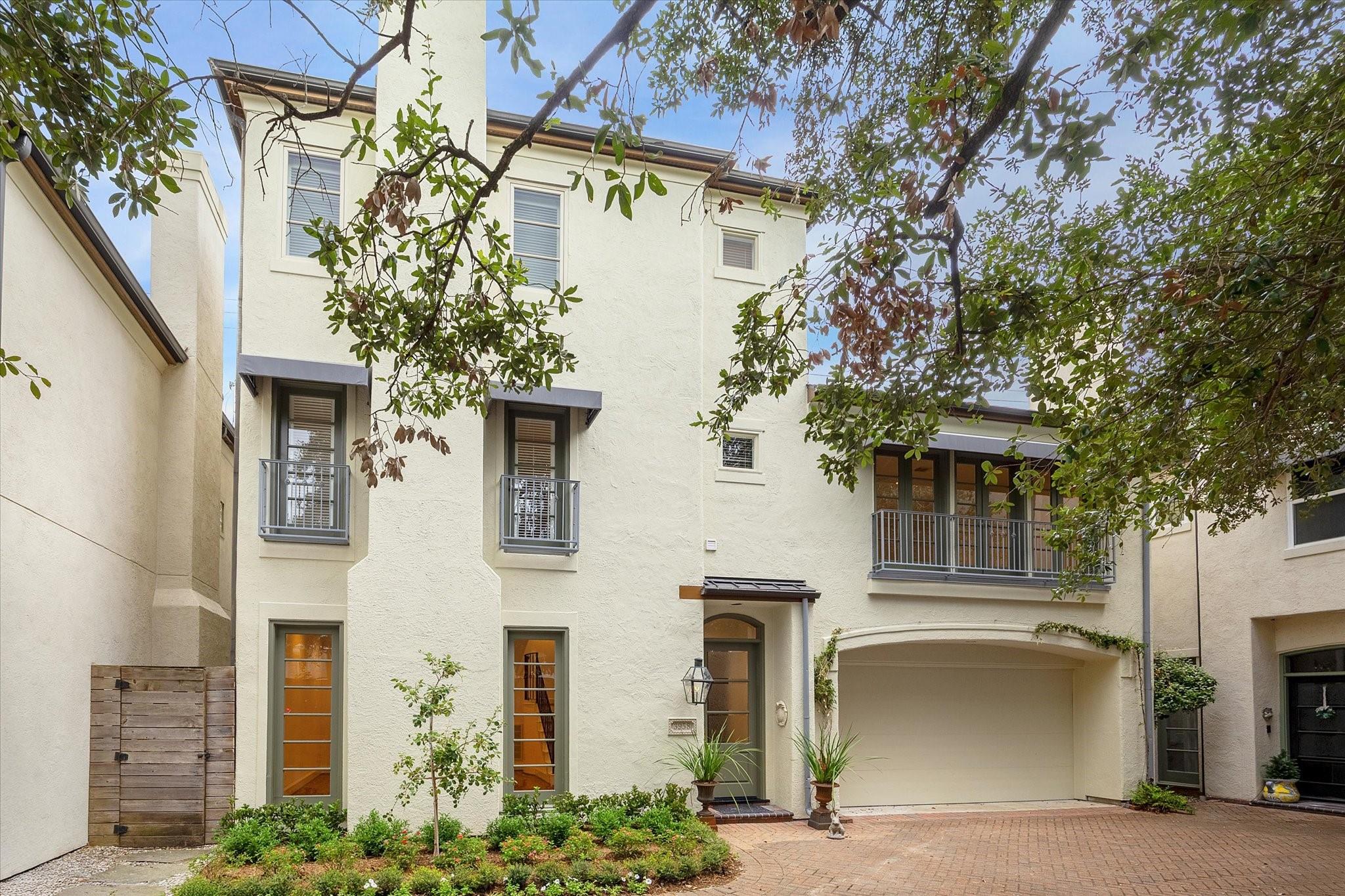 Cream stucco townhouse with balconies, an attached garage, and a tree-shaded brick courtyard.
