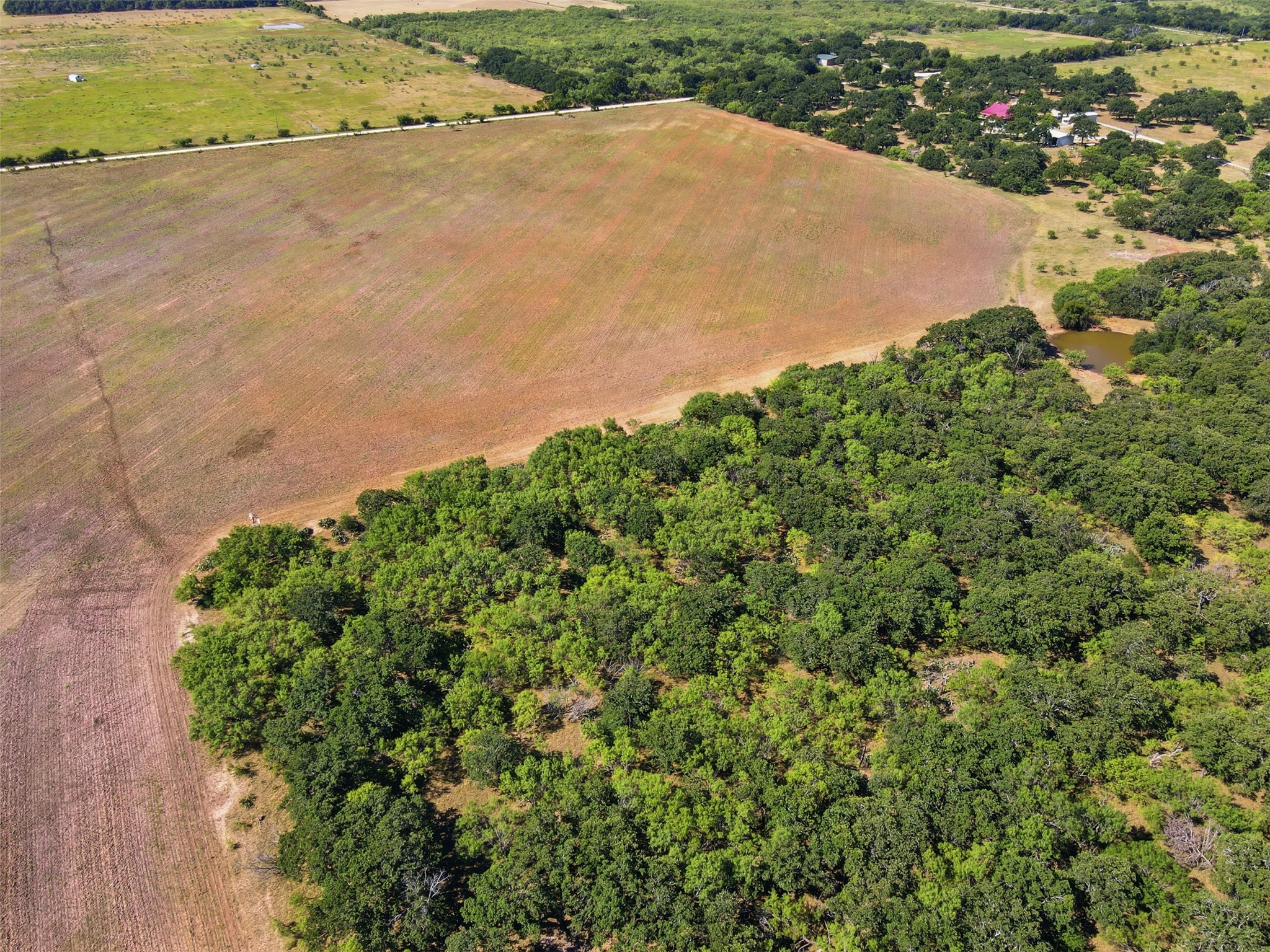 Aerial view of a plowed field bordered by dense woodland, with a small pond nearby.