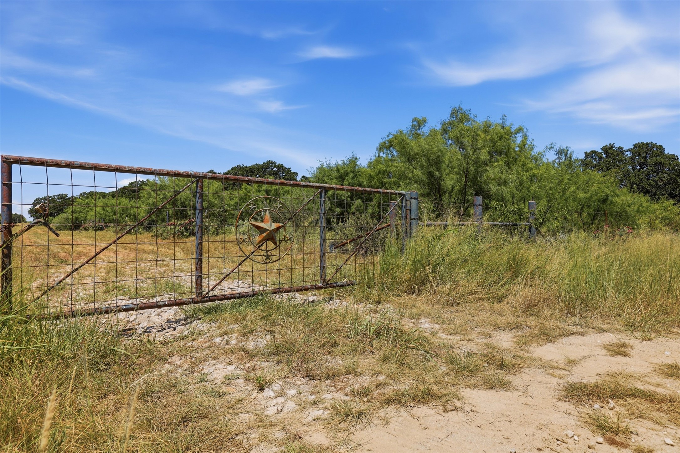 Rural property entrance with a rusted star-emblem gate, dry grass, and trees.