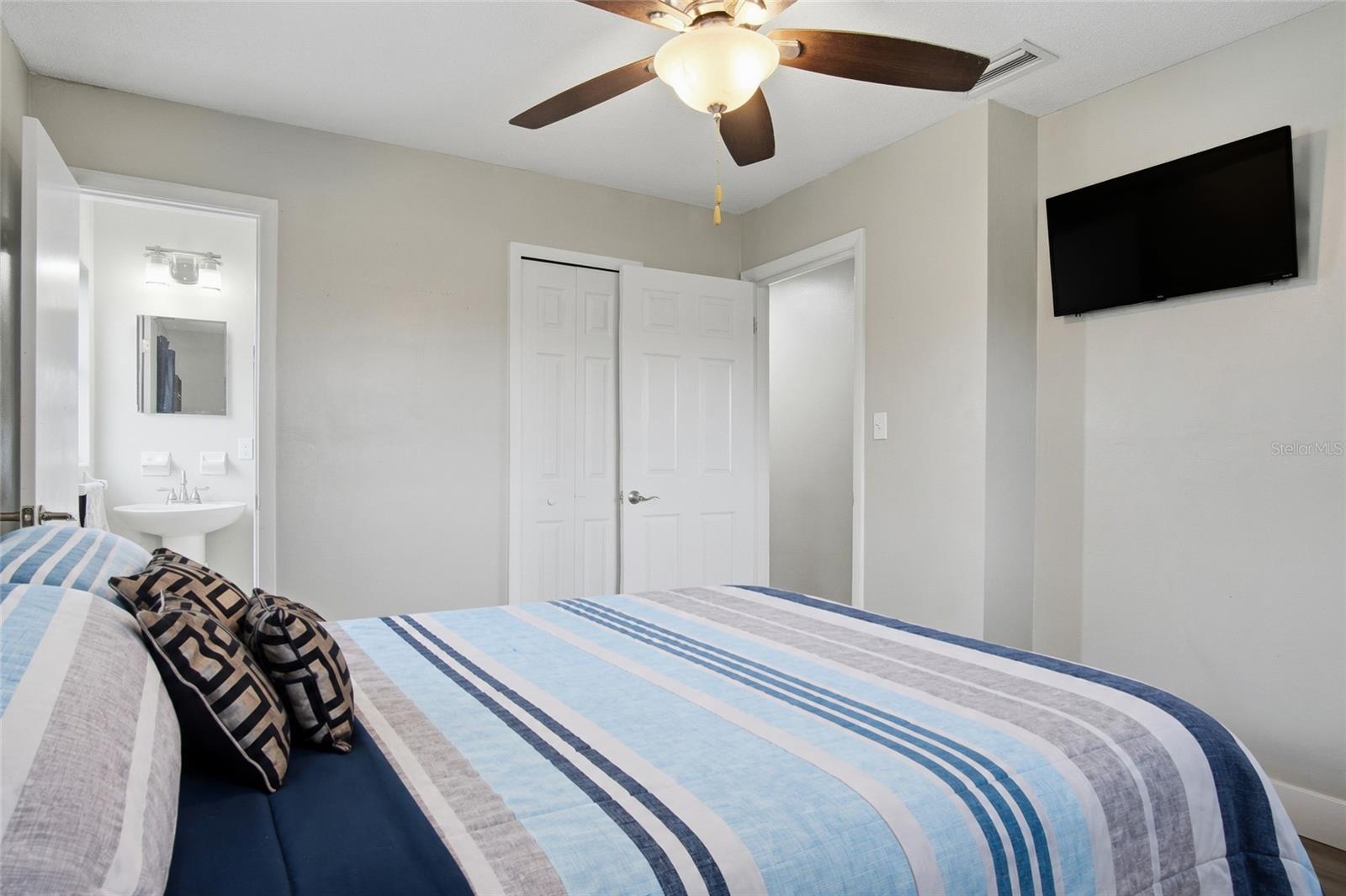 Bedroom with striped blue bedding, a ceiling fan, wall-mounted television, and bathroom vanity visible through the doorway.