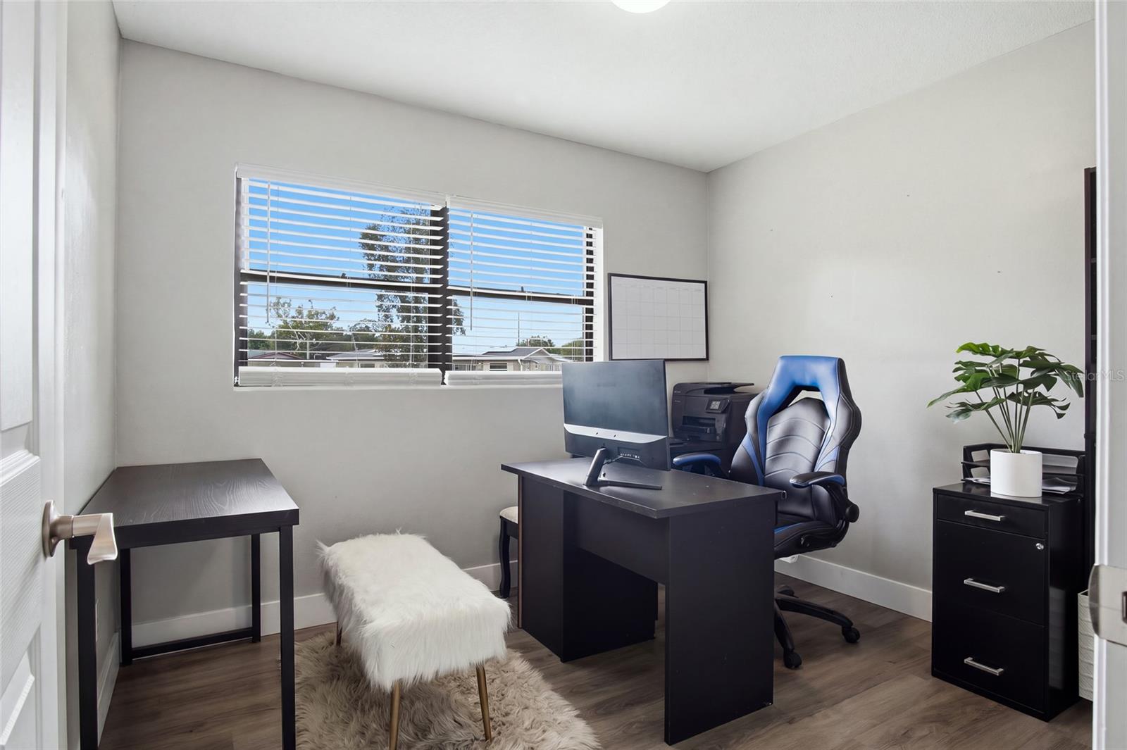 Furnished home office with dark desks, a blue office chair, and a wide window with blinds.