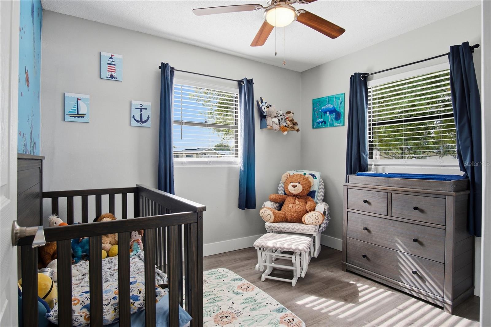 Furnished nursery with a dark wooden crib, gray dresser, teddy bear, and blue curtains.